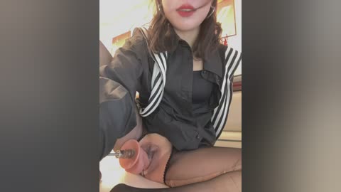 Media: Video of a woman with fair skin, shoulder-length brown hair, and red lipstick, wearing a black jacket with white stripes, sitting in a dimly lit room, holding a dildo.