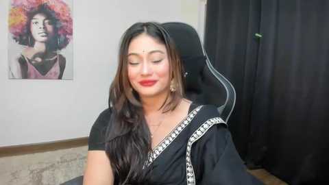 Media: Video of a woman with long, straight black hair, wearing a black sari with white border, sitting on a black chair. Background includes a colorful painting of a woman with a large afro, and black curtains.