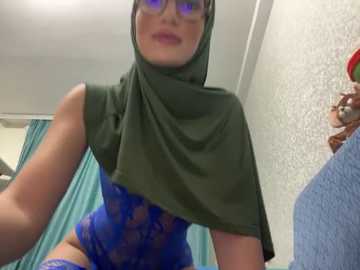 Media: Video of a fair-skinned woman with a medium build, wearing a green hijab and a revealing blue lace bodysuit, standing in a hospital room with a patterned wall and teal curtains.