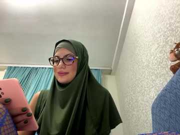 Media: Video of a light-skinned woman with glasses, wearing a green hijab and a sleeveless top, taking a selfie in a room with white textured walls and teal curtains.