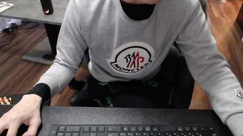 Media: A video of a person wearing a grey sweatshirt with a logo, black long sleeves, and black pants, typing on a black laptop on a wooden floor.