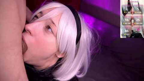 Video of a young, pale-skinned woman with platinum blonde hair and a black headband, performing oral sex on a man, set in a dimly lit room with purple lighting.