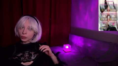 Media: A video of a pale-skinned, androgynous person with platinum blonde bobbed hair, wearing a black lace top, and black headphones. They are seated in a dimly lit room with red curtains and a purple glow from a video game screen.