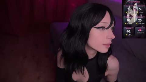 A video of a pale-skinned, slender woman with long black hair and black eyeliner, wearing a black top, sitting against a dark background. The image includes a digital overlay displaying her social media stats.