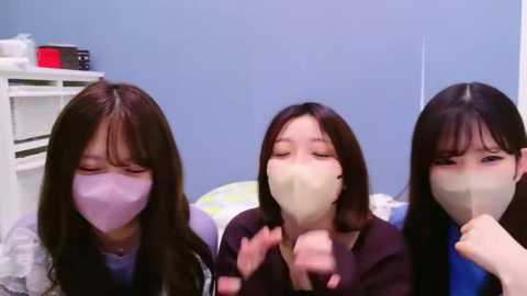 Media: Video of three Asian women wearing face masks, smiling and holding their fingers up in a \"V\" gesture, against a blue wall with a white shelf and medical supplies.