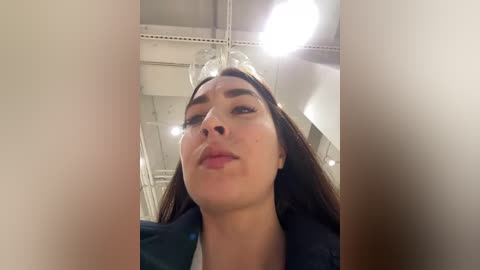 A video of a woman with long brown hair and light skin, wearing glasses and a dark blue jacket, taken from a low angle, indoors with white ceiling and lighting.