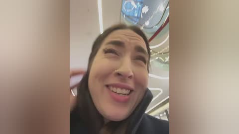 A video shows a woman with long brown hair and a broad smile, wearing a black coat, captured from a low angle, inside a subway car with reflective surfaces and blue lighting.