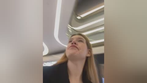 Media: Video of a young Caucasian woman with light skin and shoulder-length blonde hair, wearing a black jacket, gazing upward in a modern, well-lit interior with curved white ceiling lights.