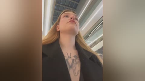 Media: Video of a Caucasian woman with long, blonde hair, wearing a black blazer, standing in a modern building with a glass ceiling and metal beams.