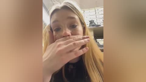 Media: Video of a blonde woman with long hair, covering her mouth with her hands, sitting in an office with shelves filled with boxes.