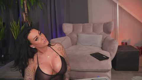 Media: Video of a curvaceous woman with long black hair and tattoos, wearing a sheer black halter top, sitting in a modern living room with a beige armchair and a laptop.