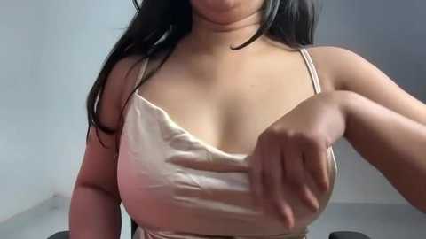 Media: Video of a young woman with medium skin tone, long black hair, wearing a satin, light pink camisole, adjusting her bra strap. Background is a plain, light-colored wall.