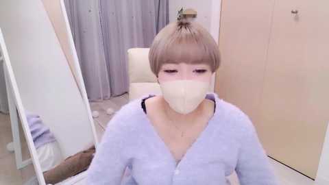 Media: Video of a young Asian woman with short, light brown hair, wearing a white face mask, a light blue cardigan, and a black top, seated in a modern, clean room with white walls and beige curtains.