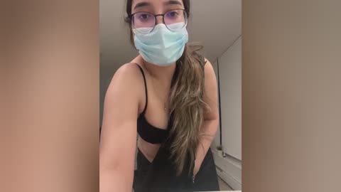 Media: Video of a young woman with long brown hair and a light complexion, wearing a blue surgical mask, black bra, and pants, leaning forward in an indoor setting.