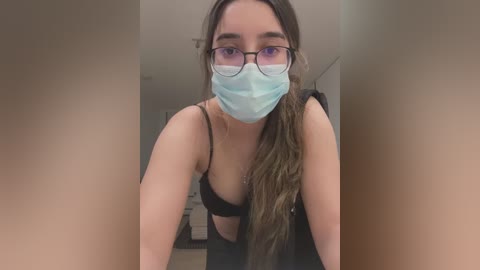 Media: Video of a young woman with light skin, glasses, and a blue surgical mask, wearing a black bra, leaning forward. Background shows a dimly lit room with a door and a white shelf.