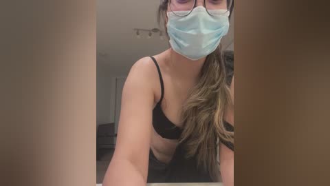 Media: Video of a woman with light brown skin and long brown hair, wearing a black bra and blue mask, captured from a low angle in a dimly lit room with beige walls.