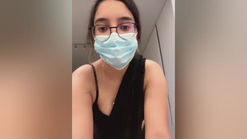 Media: Video of a young woman with light skin, wearing a blue face mask, black glasses, and a black tank top, standing in a beige hallway with a white door.