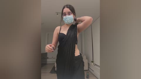 Media: A video of a young woman with light skin, dark hair, wearing a black, sleeveless dress, blue face mask, and glasses, standing in a modern hallway with beige walls.