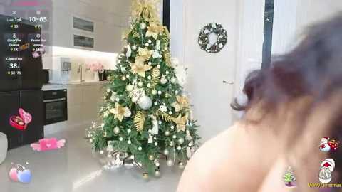 Media: Video of a woman with wavy brown hair, topless, standing in front of a decorated Christmas tree in a modern kitchen, with festive decor and digital clock in the background.