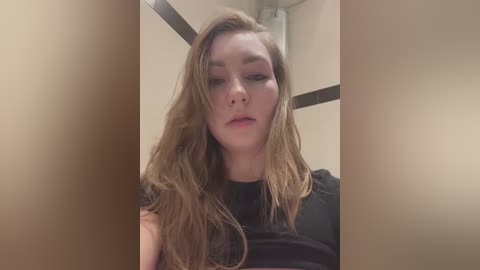 Media: Video of a young Caucasian woman with long, wavy, light brown hair, wearing a black top. She has a neutral expression. Background features beige walls and a dark horizontal line.