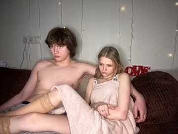 Media: Video of a young, shirtless, Caucasian boy with short brown hair and a girl with long blonde hair, both sitting on a dark brown couch in a dimly lit room with string lights and a red and white blanket.