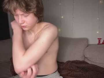 Media: Video of a young, shirtless boy with fair skin and brown hair, hugging himself in distress, seated on a beige couch. The background features white string lights and a red toy block.