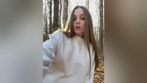 Media: Video of a young woman with long, light brown hair, wearing a white hoodie, standing in a forest with bare trees and autumn leaves.