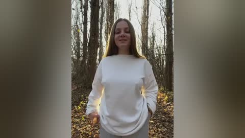 Media: Video of a young Caucasian woman with long, straight, light brown hair, wearing a white long-sleeve shirt and grey pants, standing in a forest with bare trees and fallen leaves, smiling softly.