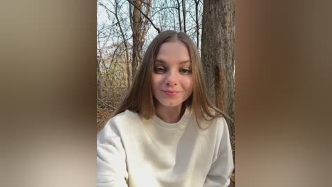 Media: Video of a young woman with long brown hair, wearing a white sweater, standing in a forest with bare trees and autumn leaves.