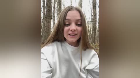 Media: Video of a young Caucasian woman with long, straight, light brown hair and fair skin, wearing a white sweatshirt, standing in a forest with bare trees and an overcast sky.