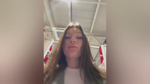 Media: Video of a young woman with long, light brown hair, fair skin, and minimal makeup, wearing a white top, standing in an industrial room with exposed pipes and red and white flags in the background.