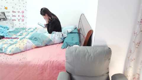 Media: Video of a young Asian woman with long black hair, wearing a black top, sitting on a pink bed with blue bedding, reading a book, in a minimalist room with white walls.