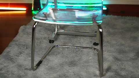 Media: A video of a transparent, neon-lit chair with metallic legs on a plush, gray rug, set against a modern, dimly lit background.