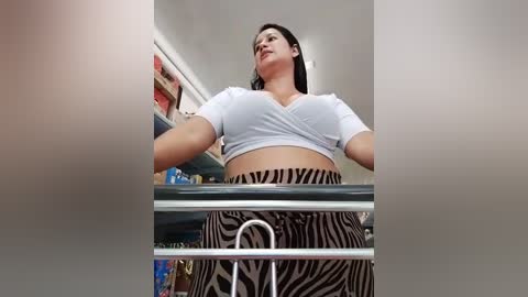 Media: Video of a woman with medium skin tone and long black hair, wearing a white cropped top and zebra-patterned pants, standing in a modern kitchen with stainless steel appliances and white countertops.