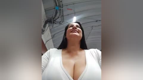 Video of a curvy woman with large breasts, wearing a low-cut white top, taken from a low angle, showing her cleavage prominently. She has long dark hair and is smiling. Background includes industrial elements like exposed pipes and cables.