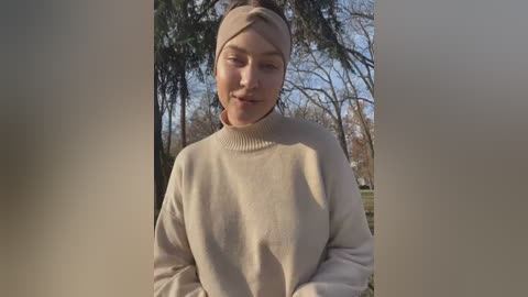 Media: A video of a young woman with light skin and brown hair in a beige sweater, standing in a sunny park with leafless trees.