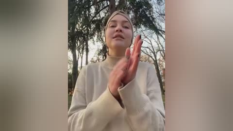 Media: Video of a woman with light skin, wearing a cream-colored turtleneck sweater and beige headband, clapping her hands in front of a forest background with tall trees and greenery.