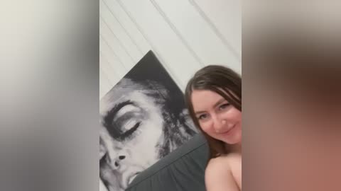 Media: Video of a smiling, light-skinned woman with shoulder-length brown hair, partially nude, standing in front of a black-and-white portrait of a woman with closed eyes on a white wall.