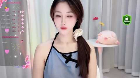 Media: Video of a young Asian woman with pale skin, brown hair tied in a ponytail, wearing a sheer blue apron. Background features a table with a pink stuffed toy and a green \"DINGA\" logo.