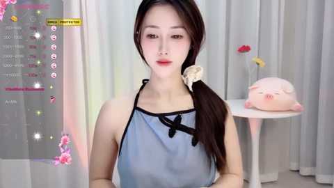 Media: Video of an East Asian woman with fair skin and long dark hair, wearing a light blue apron, standing in a minimalist room with white curtains and a pink piggy bank.