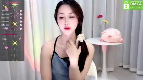 Media: A video of a young East Asian woman with fair skin, long black hair, and red lipstick, wearing a denim apron and black tank top, sitting in a minimalist room with white curtains and a pink pig pillow.