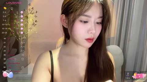 Media: Video of an East Asian woman with long brown hair, fair skin, and red lipstick, wearing a black spaghetti strap top, looking introspective. Background features a beige wall and a window with sheer curtains.