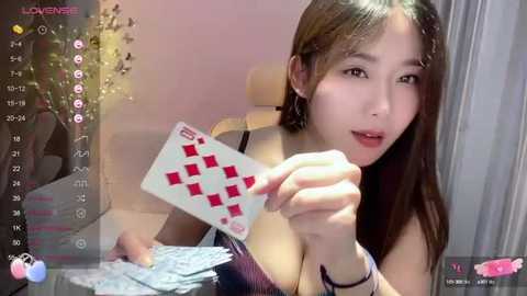 Media: Video of an Asian woman with long black hair, wearing a black top, holding a red and white playing card against a pink background.