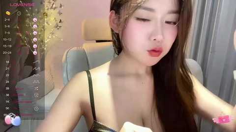 Media: Video of an Asian woman with fair skin and long brown hair, wearing a green spaghetti strap top, pouting with eyes closed, in a modern indoor setting.