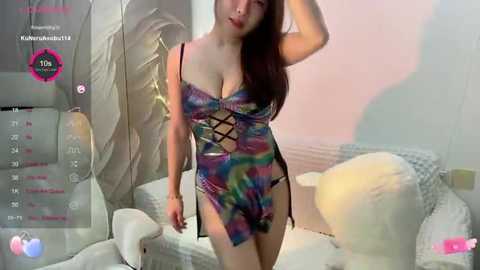 Media: Video of an East Asian woman with long brown hair, wearing a colorful, tie-dye camisole with a lacy cutout, posing in a modern, minimalist room with white furniture and soft lighting.