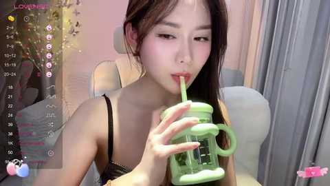 Media: A young Asian woman with long black hair sips from a green straw in a livestream setting, wearing a black bra. Background features a Christmas tree and curtains.