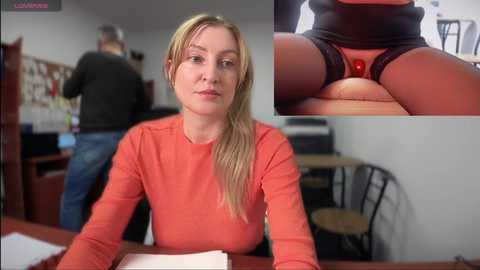 A video of a blonde woman in an orange shirt seated at a desk, with a close-up of her crotch in red lace panties. Background shows a man in a black shirt and jeans.