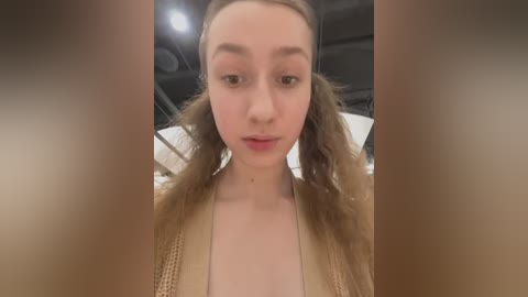 Media: Video of a fair-skinned young woman with long, wavy brown hair styled into pigtails, wearing a beige knit cardigan, indoors with industrial ceiling lights.