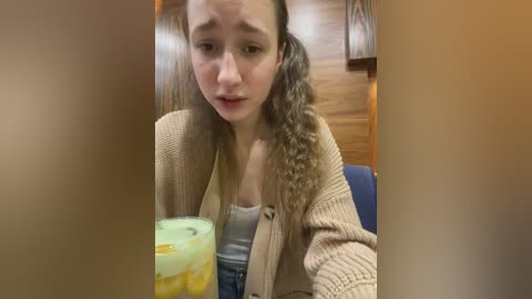 Media: Video of a young woman with long, wavy brown hair in pigtails, wearing a beige cardigan over a gray tank top and denim shorts. She holds a green plastic cup with yellow liquid, in a dimly lit room with wooden paneling.