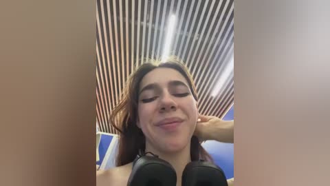 Media: Video of a young woman with light skin, long brown hair, and closed eyes, resting her head on black headphones, in a modern room with striped ceiling lights and blue accents.